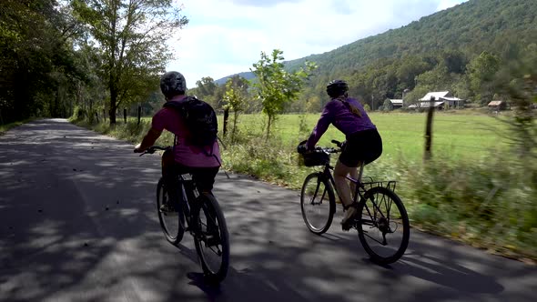 Camera following behind a mother and teenage son biking side by side on a rural road with a farm in