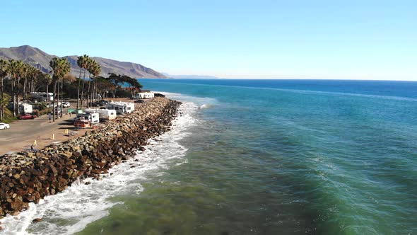 Aerial drone shot rising up over ocean waves crashing on the rocky coast with palm trees and rv camp