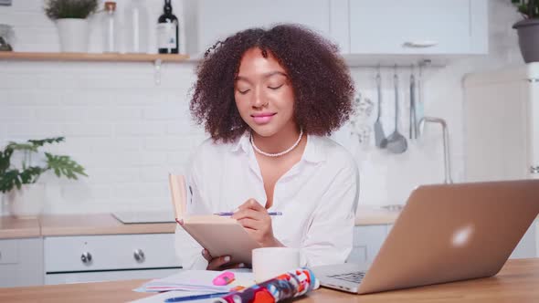 Successful African American Female Taking Notes in Workbook Looking at Laptop