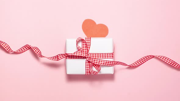 White gift box with a red ribbon bow and red hearts on pink background. Flat lay top view stop motio alt