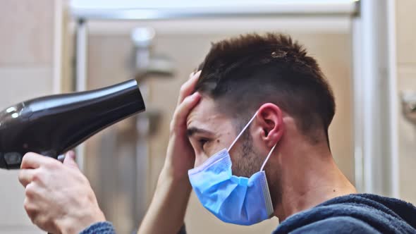 Man blow dry his hair in the bathroom wearing protective medical mask.