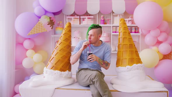 Young man with dreadlocks drinking refreshing soda, sitting on table in confectionery.