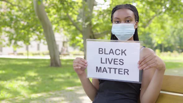 A Young Black Woman in a Face Mask Shows a Black Lives Matter Sign To the Camera and Nods