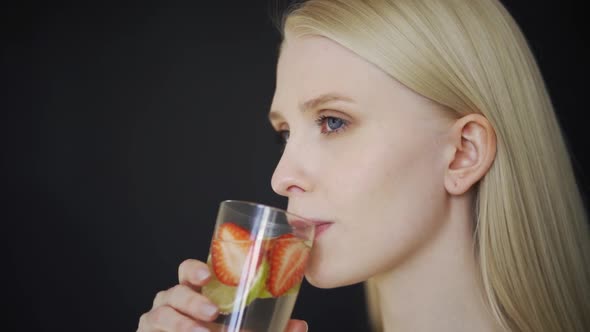 Young Blonde Woman Drinks Water with Strawberries and Lime on a Black Background Closeup