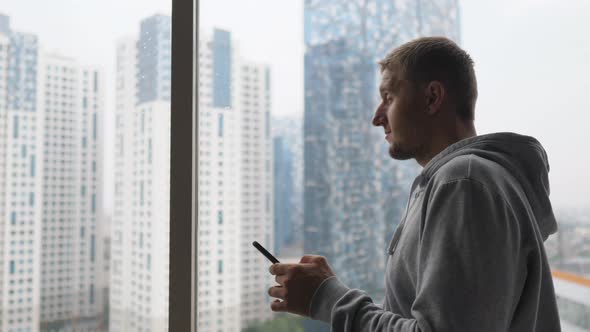 Locked in an Apartment Man Stands By the Window with His Phone Dreaming About Go Outside After
