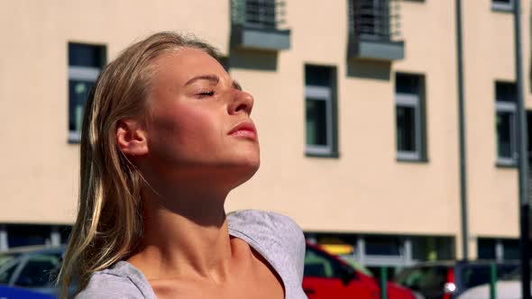 Young Pretty Blond Woman Sits on the Bench and Sunbathes in Front of Building - Closeup of Face