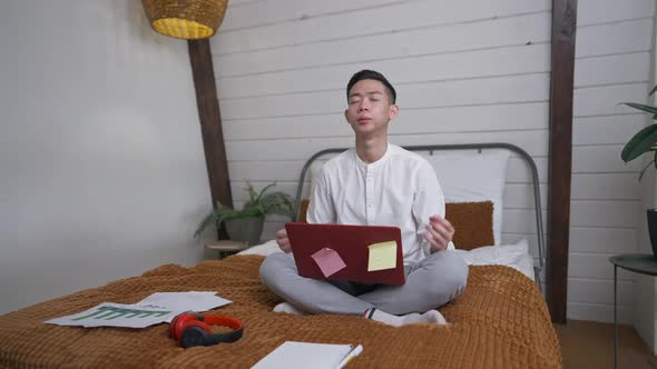 Tired Young Asian Man Sighing and Sitting on Bed in Lotus Pose Calming Down