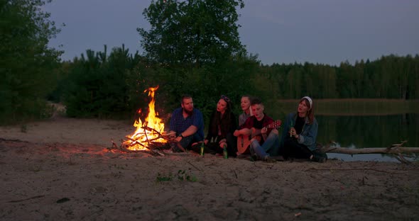 Adult Diverse Teenagers Gathering Around Burning Bonfire in Campsite and Singing Sitting in Twilight