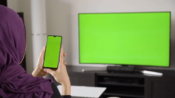 A Muslim Woman Looks at a Smartphone and TV (Green Screen) As She Sits on a Sofa in an Apartment