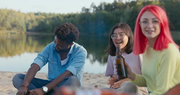 A Group of Friends is Sitting on the Beach By the Lake Drinking Beer By the Fire Singing Songs