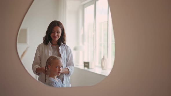 Beautiful Smiling Girl with Blond Curly Hair Looking at Her Reflection in Mirror While Her Mother