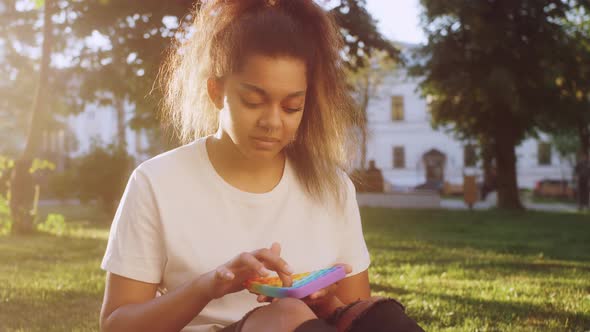Mixed Race Teenage Girl Sitting on a Grass at the Street and Holding POP IT Fidget Toy Antistress in
