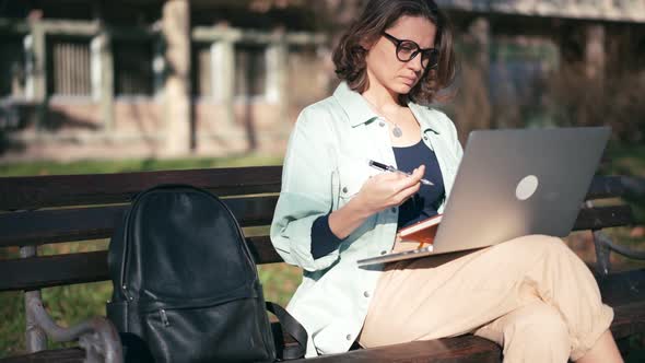 A Young Woman Student Sitting on a Bench in the Park and Typing on Her Laptop