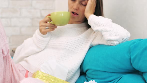 Sick Young Woman Sits of Sofa and Drinks Hot Beverage at Home