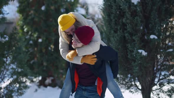 Cheerful Girlfriend Jumping on the Back of Boyfriend Posing Outdoors on Sunny Winter Day