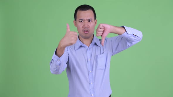 Confused Asian Businessman Choosing Between Thumbs Up and Thumbs Down