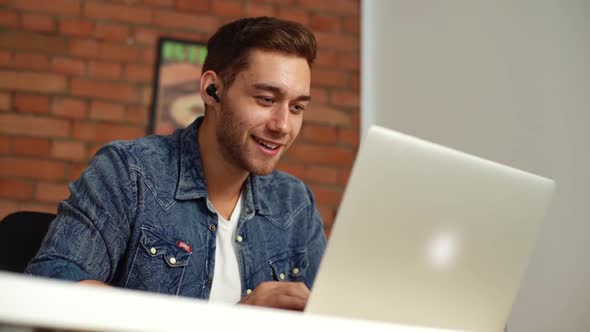 Closeup Lowangle View of Cheerful Young Man in Headphones Typing on Laptop Keyboard and Talking