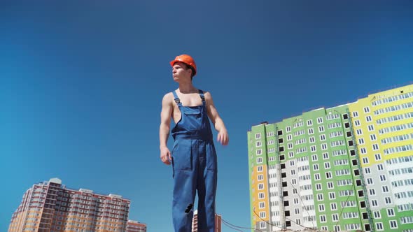 Serious Male Builder in Work Uniform and Hardhat Background of Buildings Under Construction.