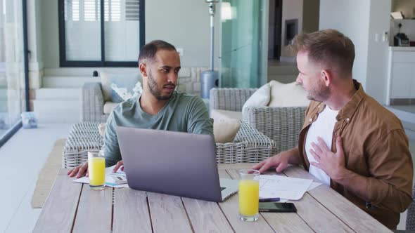 Multi ethnic gay male couple sitting at table at homw using laptop