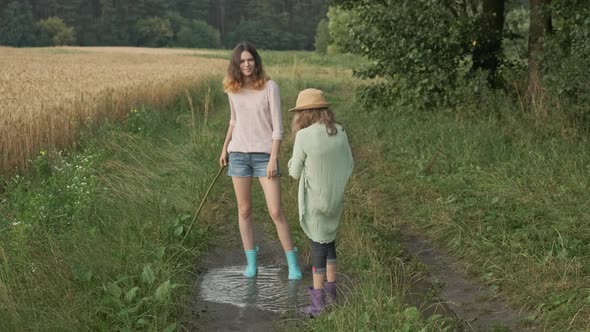 Two Happy Girls Sisters in Boots Playing in Puddle of Rainwater