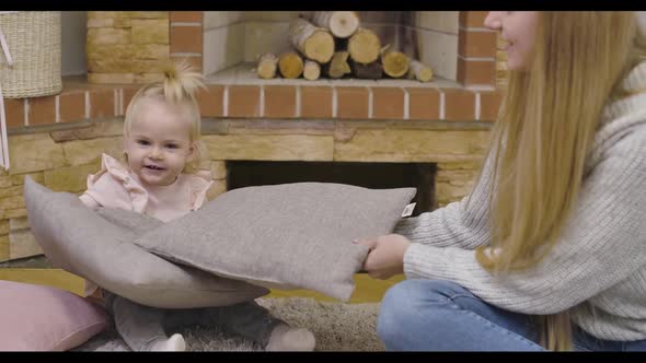 Charming Little Caucasian Girl Sitting with Mother on Soft Carpet and Playing with Pillows