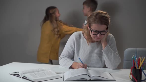 Exhausted Caucasian Mother in Eyeglasses Sitting at the Table and Doing Homework for Her Children