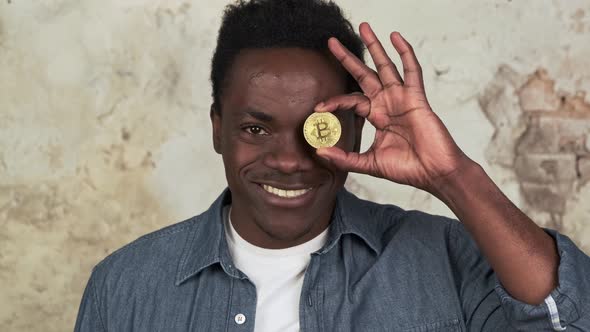 Funny African American Man with Charming Smile Standing Covering One Eye with Golden Bitcoin Closeup