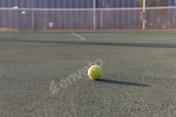 yellow tennis ball sits on a green tennis court, with a blurred background of a tennis net and trees