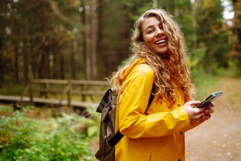 Travel apps. Curly young woman traveler in yellow coat holding smartphone in hand. Concept of travel