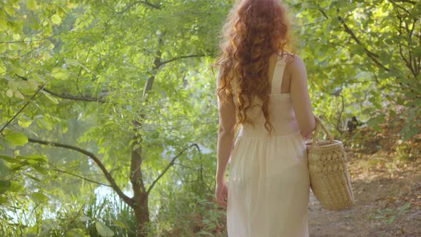 Back View of Beautiful Slim Woman with Red Hair Walking in the Garden or Park Holding Basket