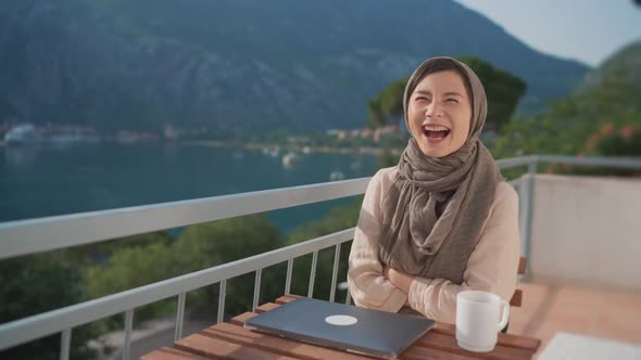 Fun Arab Young Muslim Woman in Hijab Laughing at Camera Sits Balcony Using Laptop Working Remote