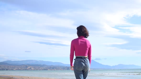 Dressed Young Woman Goes Along the Coastline on the Mountain Background