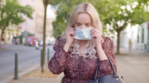 A Middleaged Caucasian Woman Puts on a Face Mask and Looks at the Camera in an Urban Area