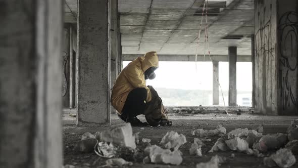 Nervous Man in Yellow Protective Suit and Gas Mask Squats in Abandoned Building Takes Out Child Toy