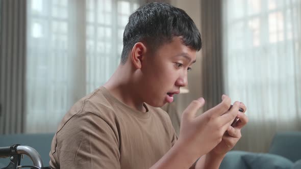 Close Up Excited Young Asian Boy Sitting On Wheelchair Playing Video Game On Mobile Phone In House