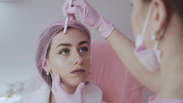 Young Woman Doctor Beautician in White Lab Coat and Sterile Gloves Draws a Marking on Woman's