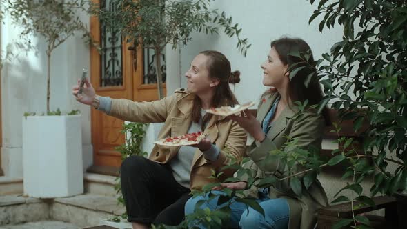 Two woman eating  pizza at the street, they are taking pictures of food with their smartphones