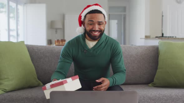 Man wearing Santa hat opening gift box while having video chat on his laptop