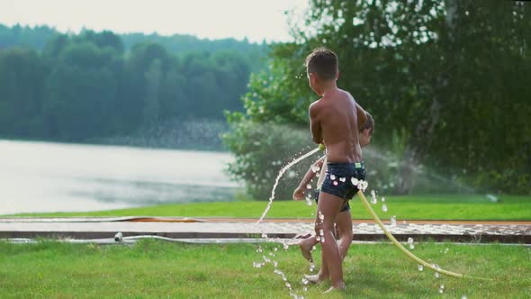 Boy in Summer Swimming Trunks Pours Water on His Younger Brother Having Fun in the Park on the Grass