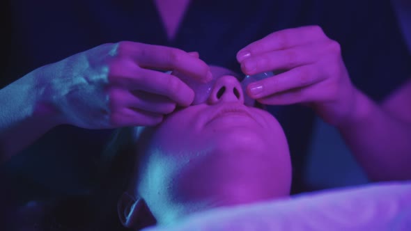 Woman Massagist Doing a Face Massage Using Small Jade Pieces on Her Female Client  Neon Lighting