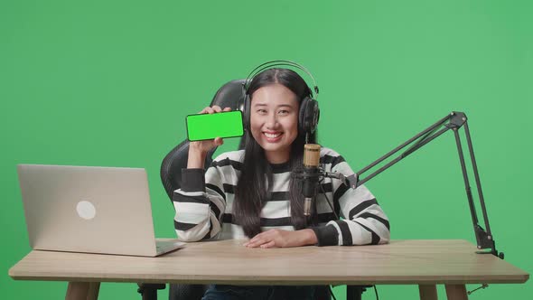 Asian Woman With Headphone Showing Green Screen Mobile Phone While Sitting In Front Of Green Screen