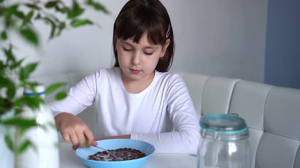 Little Girl Eats Chocolate Corn Balls with Milk Sitting at White Table