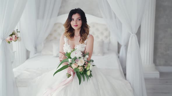 Attractive young bride in a sexy wedding dress is sitting on bed. Beautiful bride with flowers.