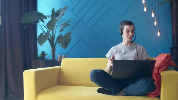 Woman with Short Hair and a Headset is Talking on Video Link a Laptop While Sitting on Yellow Sofa