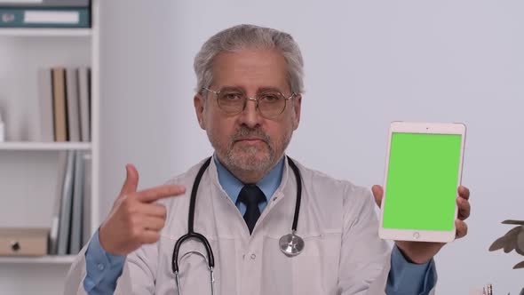 Aged Male Doctor Holds Tablet with Green Screen Chroma Key Points at It with Finger and Makes Thumb