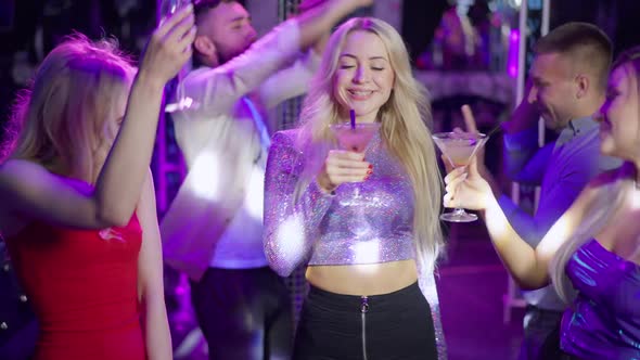 Gorgeous Young Caucasian Women Clinking Glasses Drinking Cocktails As Men Dancing at Background