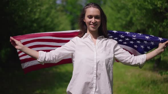 Portrait of Proud Slim Young Woman Wrapping in US Flag Looking at Camera Smiling