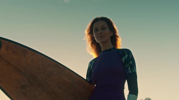 Young Attractive Female Surfer Smiling and Posing with Her Surfboard at Sunset on the Beach