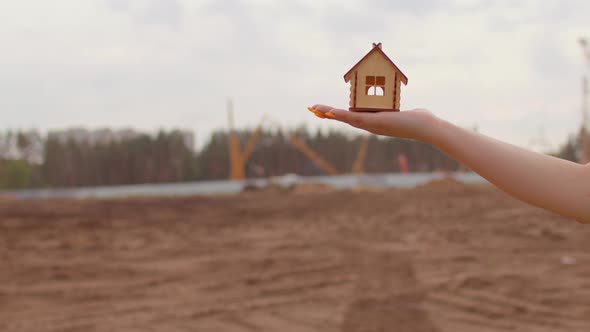 Close Up of Woman's Hand Holding Small Wooden House on Background of Construction Site