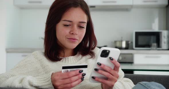 Smiling Brunette Woman Customer Holding Credit Card and Smartphone Sitting on Couch at Home